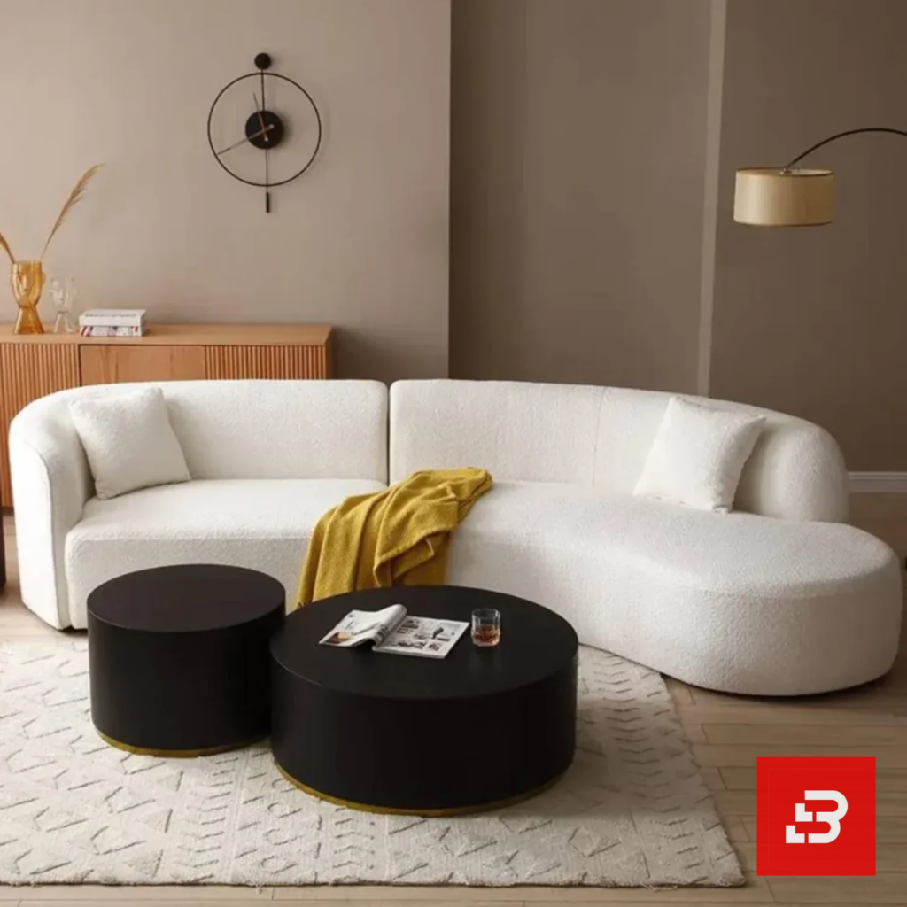 Cozy living room view of a luxury upholstered modern curved sectional sofa in off white bouclé fabric styled with accent pillows, a yellow throw blanket, and black round coffee tables by Bahoo Furniture UAE