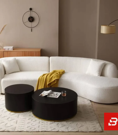 Cozy living room view of a luxury upholstered modern curved sectional sofa in off white bouclé fabric styled with accent pillows, a yellow throw blanket, and black round coffee tables by Bahoo Furniture UAE