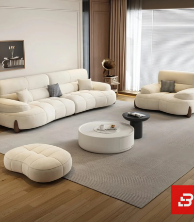 Bright modern living room view of a luxury upholstered contemporary sofa set in off white velvet fabric featuring a 3-seater sofa, matching armchair, pouf, and round nesting coffee tables by Bahoo Furniture UAE