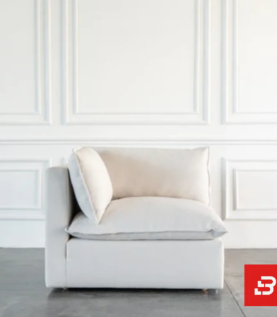 Studio interior view of a luxury upholstered modular corner lounge chair in off white velvet fabric featuring a plush back cushion and side armrest against a white wall paneling background by Bahoo Furniture UAE