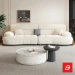 Bright modern living room view of a luxury upholstered contemporary sofa set in off white velvet fabric featuring a 3-seater sofa, matching armchair, pouf, and round nesting coffee tables by Bahoo Furniture UAE