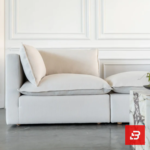 Studio interior view of a luxury upholstered modular corner lounge chair in off white velvet fabric featuring a plush back cushion and side armrest against a white wall paneling background by Bahoo Furniture UAE