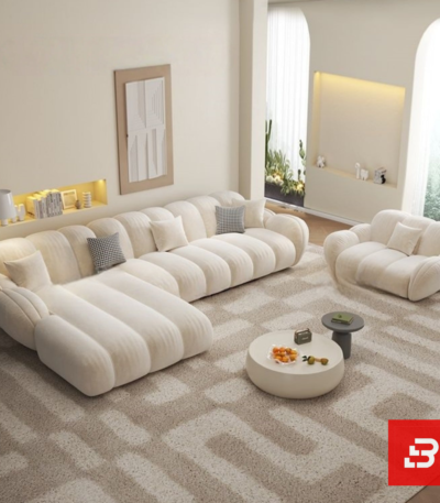 Sunlit minimal modern apartment living room interior view of a luxury channel tufted sectional upholstered in textured off white bouclé fabric featuring a generous chaise lounge and matching barrel armchair by Bahoo Furniture UAE Craftsmanship Details Tactile Comfort: Wrapped in high-density, multi-tonal bouclé that adds rich texture and subtle depth to neutral color schemes while holding up to daily lounging and heavy family use. Supportive Geometry: Every channeled cushion, extended chaise contour, and rounded armrest is padded with high-resilience foam, removing sharp angles to create a secure, comfortable, and inviting environment for relaxed evenings and effortless hosting. Low-Profile Proportions: Designed with structured low-profile silhouettes and continuous clean lines, encouraging an open, airy feeling while maintaining a commanding, sophisticated presence in the room. Sectional Fluidity: Engineered with an expansive extended chaise layout, allowing you to establish a tailored configuration that perfectly fits upscale UAE living room dimensions and open-concept floor plans. Specifications Brand: Bahoo Furniture Model: The Luxury Channel Tufted Sectional Fabric Grade: Luxury Commercial-Grade Bouclé (Off White - #F8F9FA) Internal Structure: Kiln-Dried Hardwood Frame with Reinforced Joinery Configuration Options: Extended Chaise Sectional and Matching Armchair Setup White-Glove Service: Custom Handcrafted in UAE | Complimentary In-Home Assembly Care & Preservation Routine Maintenance: Vacuum gently on low suction with a soft upholstery brush attachment. Spill Response: Blot immediately with a dry, clean microfiber cloth. Clean gentle spots using warm water and mild fabric shampoo; do not scrub forcefully.