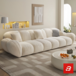 Sunlit minimal modern apartment living room interior view of a luxury channel tufted sectional upholstered in textured off white bouclé fabric featuring a generous chaise lounge and matching barrel armchair by Bahoo Furniture UAE Craftsmanship Details Tactile Comfort: Wrapped in high-density, multi-tonal bouclé that adds rich texture and subtle depth to neutral color schemes while holding up to daily lounging and heavy family use. Supportive Geometry: Every channeled cushion, extended chaise contour, and rounded armrest is padded with high-resilience foam, removing sharp angles to create a secure, comfortable, and inviting environment for relaxed evenings and effortless hosting. Low-Profile Proportions: Designed with structured low-profile silhouettes and continuous clean lines, encouraging an open, airy feeling while maintaining a commanding, sophisticated presence in the room. Sectional Fluidity: Engineered with an expansive extended chaise layout, allowing you to establish a tailored configuration that perfectly fits upscale UAE living room dimensions and open-concept floor plans. Specifications Brand: Bahoo Furniture Model: The Luxury Channel Tufted Sectional Fabric Grade: Luxury Commercial-Grade Bouclé (Off White - #F8F9FA) Internal Structure: Kiln-Dried Hardwood Frame with Reinforced Joinery Configuration Options: Extended Chaise Sectional and Matching Armchair Setup White-Glove Service: Custom Handcrafted in UAE | Complimentary In-Home Assembly Care & Preservation Routine Maintenance: Vacuum gently on low suction with a soft upholstery brush attachment. Spill Response: Blot immediately with a dry, clean microfiber cloth. Clean gentle spots using warm water and mild fabric shampoo; do not scrub forcefully.