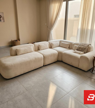 Bright natural light modern luxury living room interior view of a luxury modular sectional upholstered in textured beige bouclé fabric featuring low-profile modular corner seating, bolster pillows, and a patterned throw blanket by Bahoo Furniture UAE