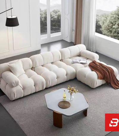 Sunlit minimal modern apartment living room interior view of a luxury segmented cushion sectional upholstered in textured off white bouclé fabric featuring pillowed modular segments and an extended chaise lounge by Bahoo Furniture UAE