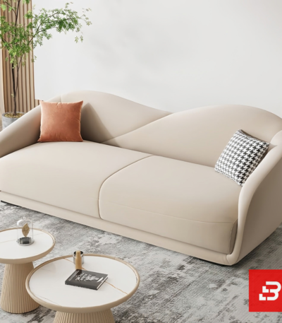 Bright modern sunlit living room interior showcasing a luxury curved sofa upholstered in smooth beige velvet fabric featuring a sweeping architectural backrest and stylish scatter cushions by Bahoo Furniture UAE