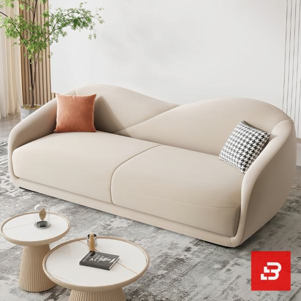 Bright modern sunlit living room interior showcasing a luxury curved sofa upholstered in smooth beige velvet fabric featuring a sweeping architectural backrest and stylish scatter cushions by Bahoo Furniture UAE
