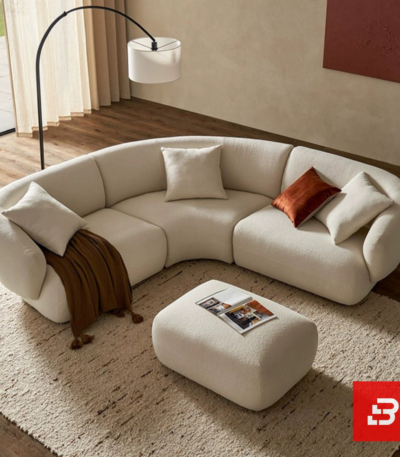 Sunlit luxury living room interior view of a luxury curved sofa upholstered in textured off white bouclé fabric featuring matching ottoman and rust accent cushions by Bahoo Furniture UAE