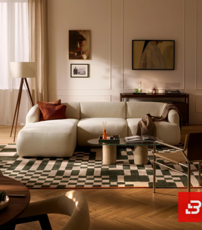 Warm modern apartment living room interior view of a luxury l-shaped sectional sofa upholstered in textured off white bouclé fabric featuring an extended chaise lounge and rust accent pillows by Bahoo Furniture UAE