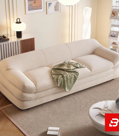 Warm sunlit modern minimalist living room interior showcasing a luxury layered sofa upholstered in textured off white bouclé fabric featuring distinctive multi-tiered horizontal cushioning and soft throw blanket by Bahoo Furniture UAE