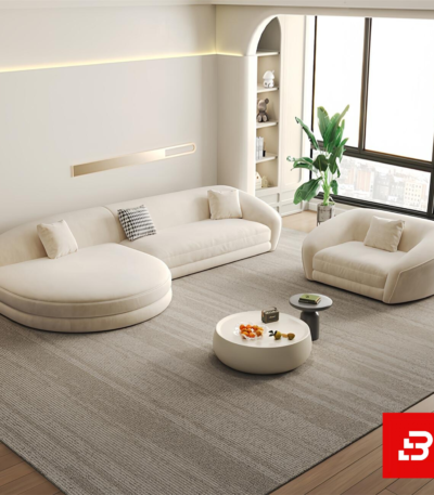 Sunlit luxury living room interior showcasing a sprawling curved chaise lounge sofa and matching armchair upholstered in textured off white bouclé fabric featuring sweeping rounded contours and a round coffee table by Bahoo Furniture UAE