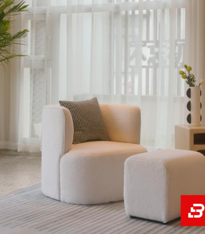 Bright modern living room interior showcasing a luxury accent armchair and matching ottoman upholstered in soft off white bouclé fabric featuring a decorative checkered throw pillow by Bahoo Furniture UAE