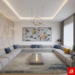 Front interior view of a luxury upholstered modern majlis sectional sofa in warm gray velvet fabric featuring blue and yellow accent pillows and a central fire feature table by Bahoo Furniture UAE