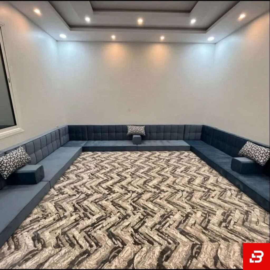 Wide interior view of a luxury upholstered floor majlis sectional sofa in warm gray velvet fabric featuring patterned accent cushions and a large abstract patterned carpet by Bahoo Furniture UAE