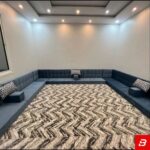 Wide interior view of a luxury upholstered floor majlis sectional sofa in warm gray velvet fabric featuring patterned accent cushions and a large abstract patterned carpet by Bahoo Furniture UAE