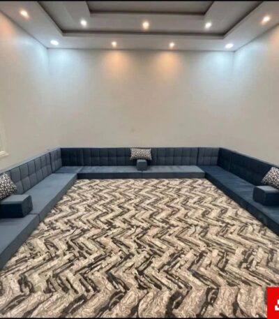 Wide interior view of a luxury upholstered floor majlis sectional sofa in warm gray velvet fabric featuring patterned accent cushions and a large abstract patterned carpet by Bahoo Furniture UAE