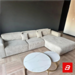 Contemporary modern living room interior showcasing a plush l-shape sectional sofa upholstered in ribbed warm gray textured corduroy paired with nested marble coffee tables against a dark accent wall by Bahoo Furniture UAE