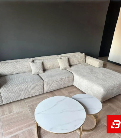 Contemporary modern living room interior showcasing a plush l-shape sectional sofa upholstered in ribbed warm gray textured corduroy paired with nested marble coffee tables against a dark accent wall by Bahoo Furniture UAE