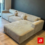 Contemporary modern living room interior showcasing a plush l-shape sectional sofa upholstered in ribbed warm gray textured corduroy paired with nested marble coffee tables against a dark accent wall by Bahoo Furniture UAE