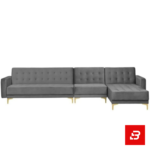 Warmly lit modern living room interior showcasing a warm gray fabric tufted l-shape sectional sofa with deep button tufting, gleaming gold metal legs, pendant lights, and indoor plant by Bahoo Furniture UAE