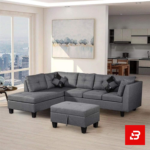 Modern open-concept apartment living room showcasing a tufted warm gray fabric l-shape sectional sofa with a matching storage ottoman and city skyline view by Bahoo Furniture UAE