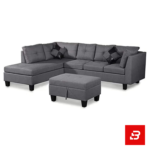 Modern open-concept apartment living room showcasing a tufted warm gray fabric l-shape sectional sofa with a matching storage ottoman and city skyline view by Bahoo Furniture UAE